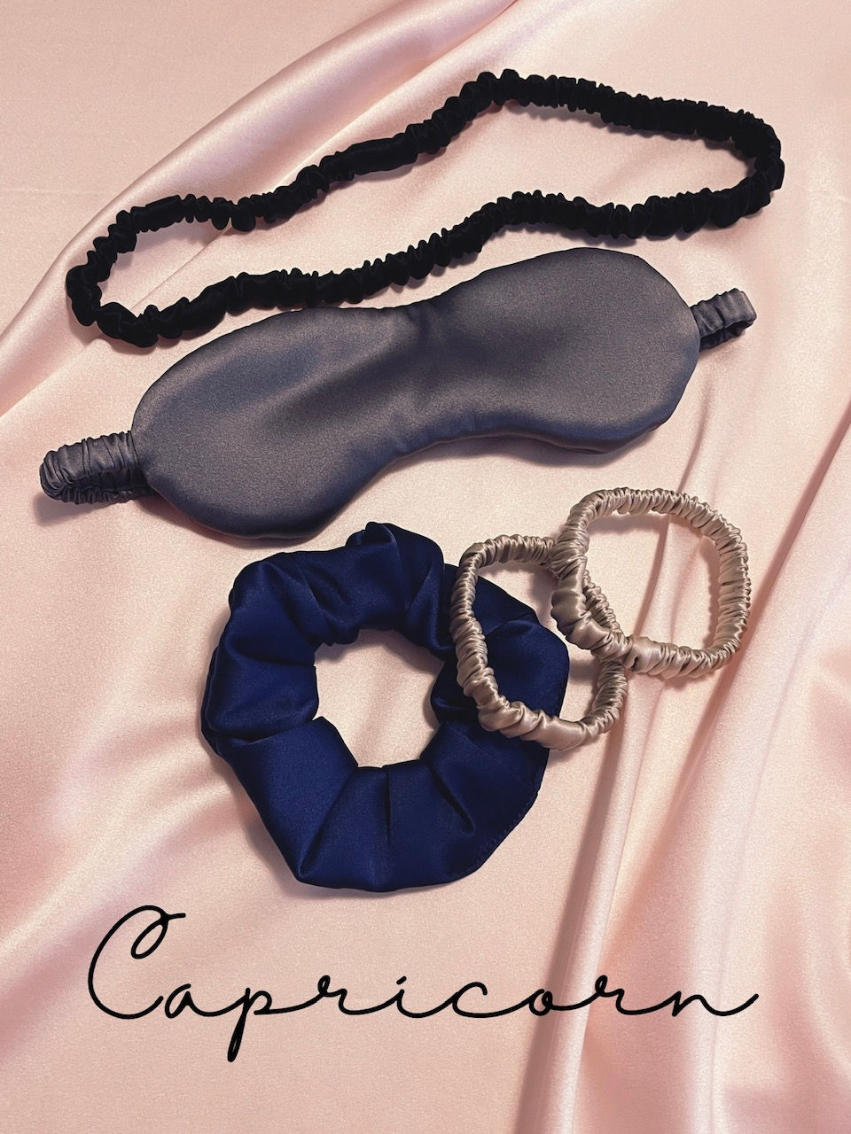 a black silk spa headband, dark grey silk eye mask, navy blue scrunchie and two golden beige silk hair elastics against a pale pink silk background with Capricorn text