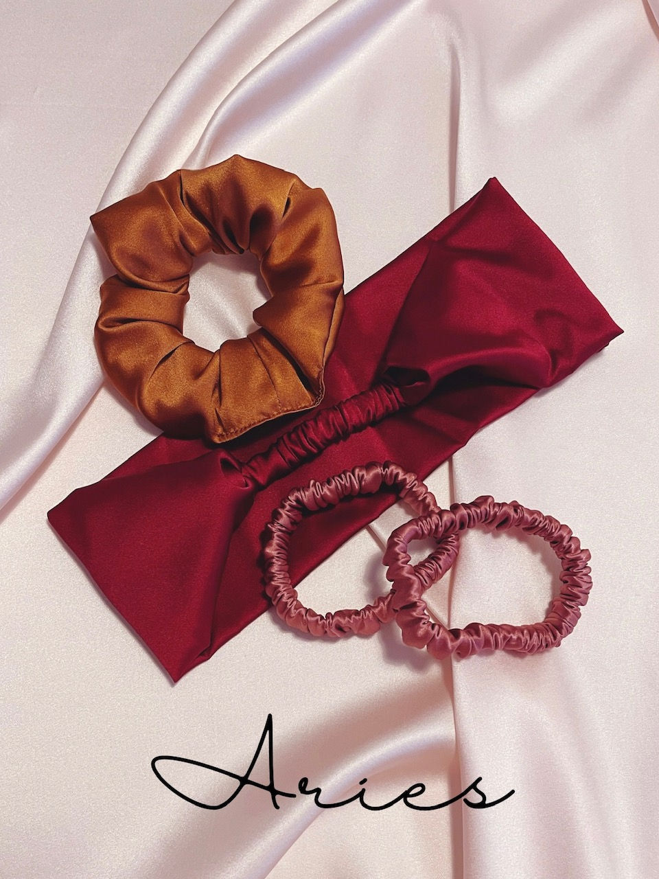 copper silk scrunchie, deep red headband, two mid pink silk hair elastics against a pale pink silk background with Aries text