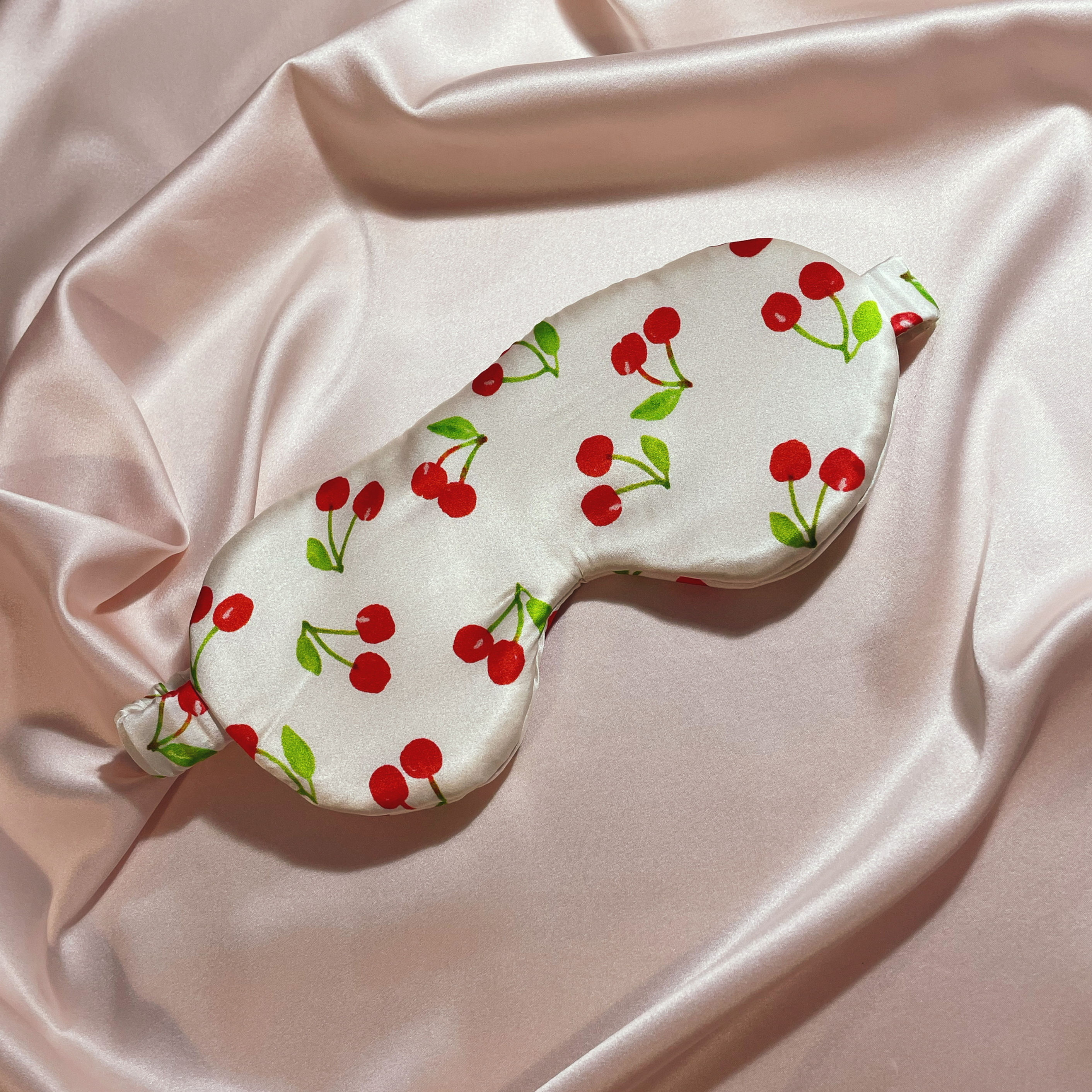 A large mulberry silk eye mask in white with a cherry print against a pale pink silk background