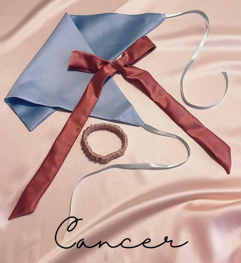 pale blue silk triangle headscarf with ribbon ties, mid pink silk ribbon and a pale pink hair elastic against a pale pink silk background with Cancer text
