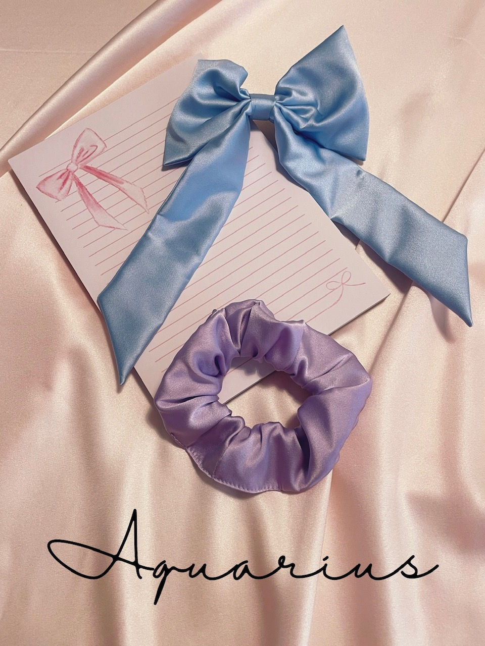 a pink lined notepad with watercolour bow details, pale blue silk hair bow and pale purple silk scrunchie against a pale pink silk background with Aquarius text