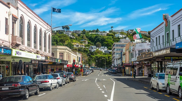 Sunlit main street with parked cars and shops, flags waving, leading to a green hillside of houses under blue sky.