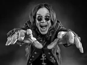 Ozzy Osbourne em apresentação ao vivo, vestindo roupas escuras e com expressão intensa, simbolizando sua icônica imagem como "príncipe das trevas" do heavy metal.