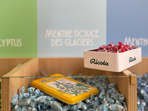 Présentoir en bois de bonbons Ricola, avec compartiments remplis de bonbons emballés bleu clair, rouges et verts, et un couvercle métallique jaune décoré de plantes au centre.