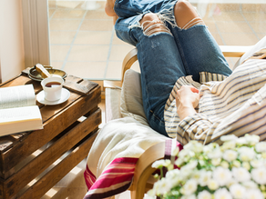 Frau entspannt in einem gemütlichen Sessel mit einem Buch, einer Tasse Tee und einer liebevoll gestalteten Umgebung mit Blumen und natürlichem Licht.
