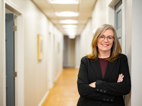 A smiling woman in professional clothing leaning up against a wall with her arms crossed in a brightly lit hallway