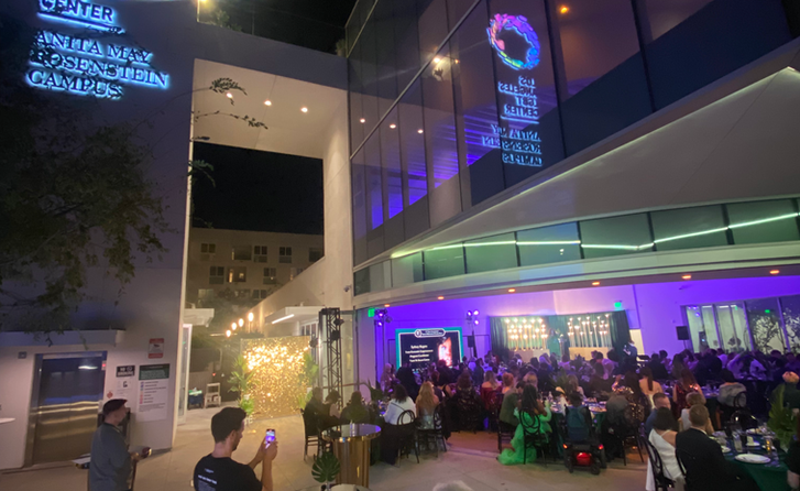 A nighttime view of an event taking place at the LA LGBT Community Center. People are standing around tables. The event was set up and run by volunteers.