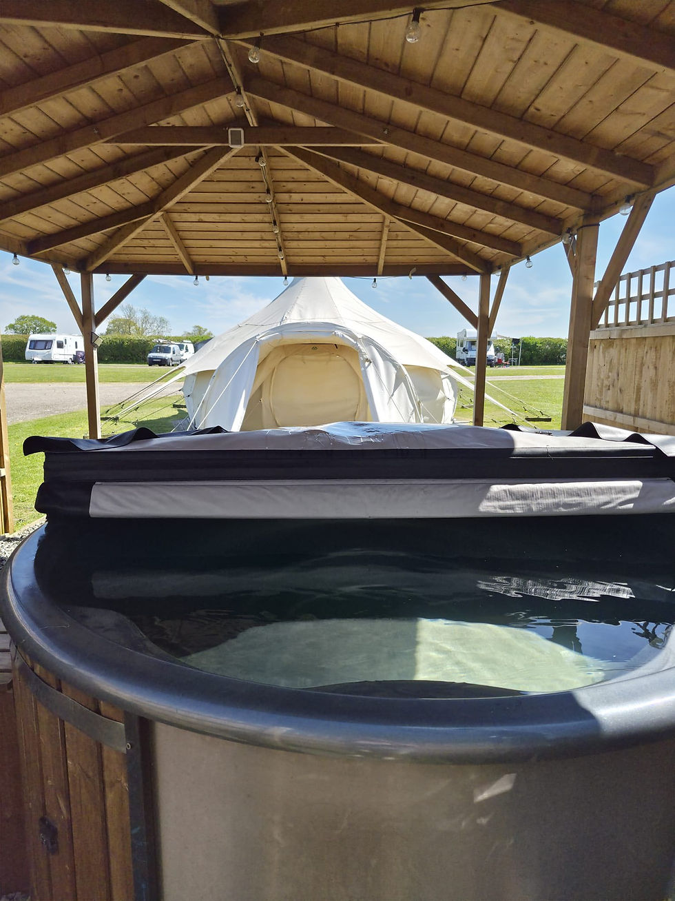 A hot tub in the foreground half uncovered under a shelter with a Hill Farm bell tent in the background underneith a wooden shelter bathed in summer sunshine.