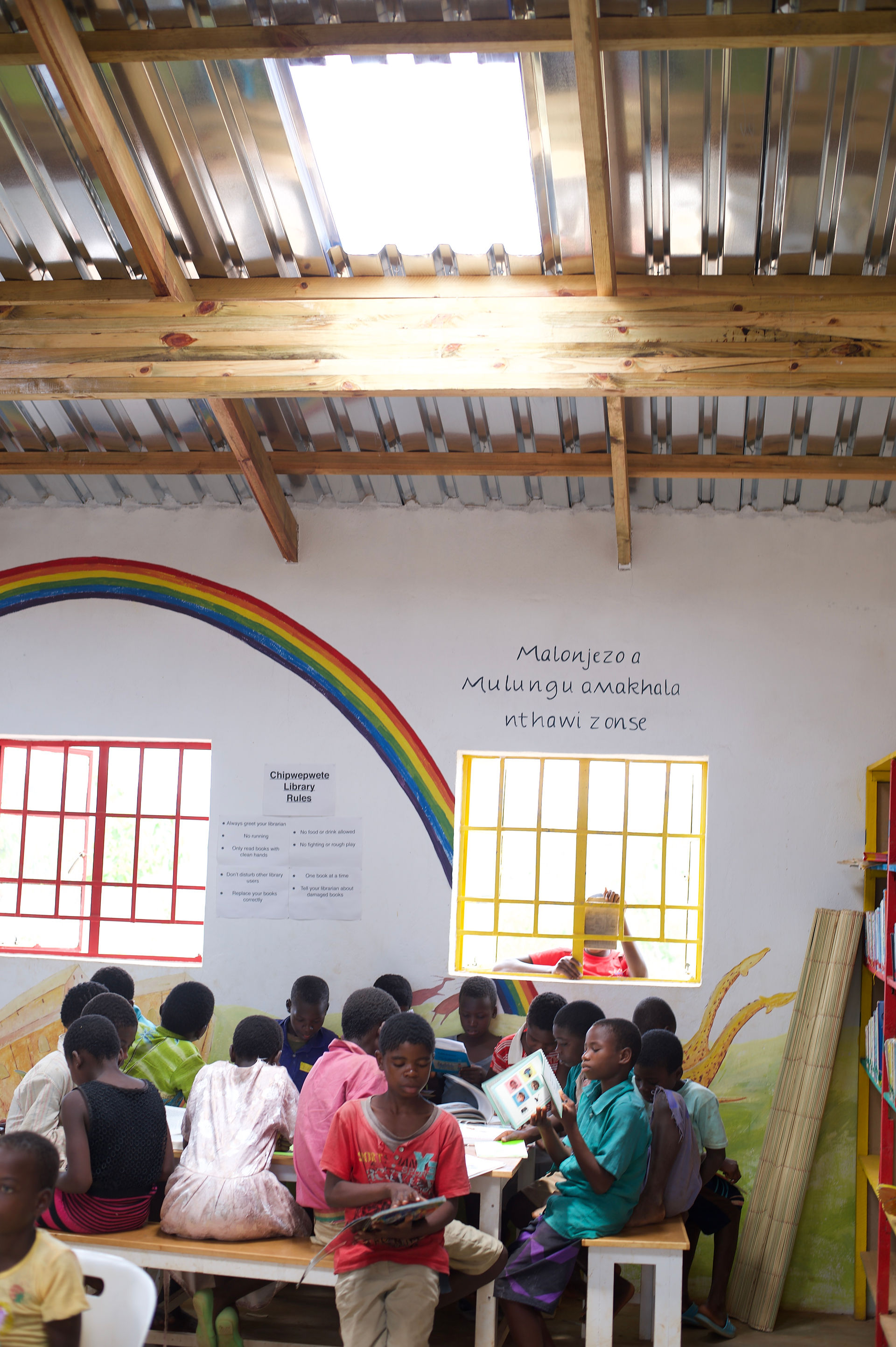 Children sitting around tables in Chipwepwete library reading books in groups. There is a rainbow painted on the wall behind them.