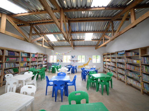 Chairs and tables laid out in groups in St Mary’s Chimwabvi Library with shelves of books on two sides of the room and a painted mural of a scene from a picture book painted on the back wall.