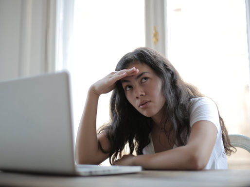 Tired woman rests her hand on her forehead while looking at a laptop by a bright window.