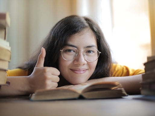 Smiling woman with glasses gives a thumbs-up behind an open book, surrounded by stacks of books in warm light.