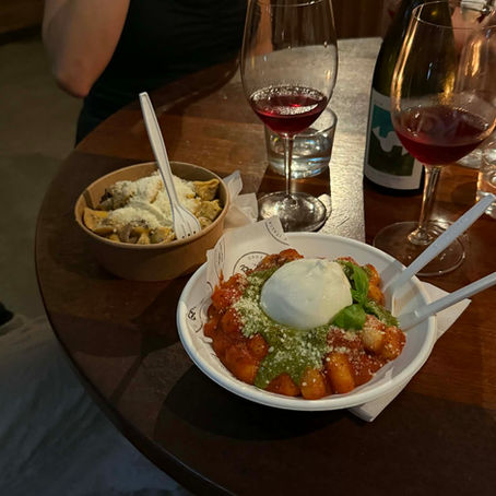 Bowl of tomato gnocchi topped with pesto and a big ball of burrata with a knife and fork, a glass of red wine
