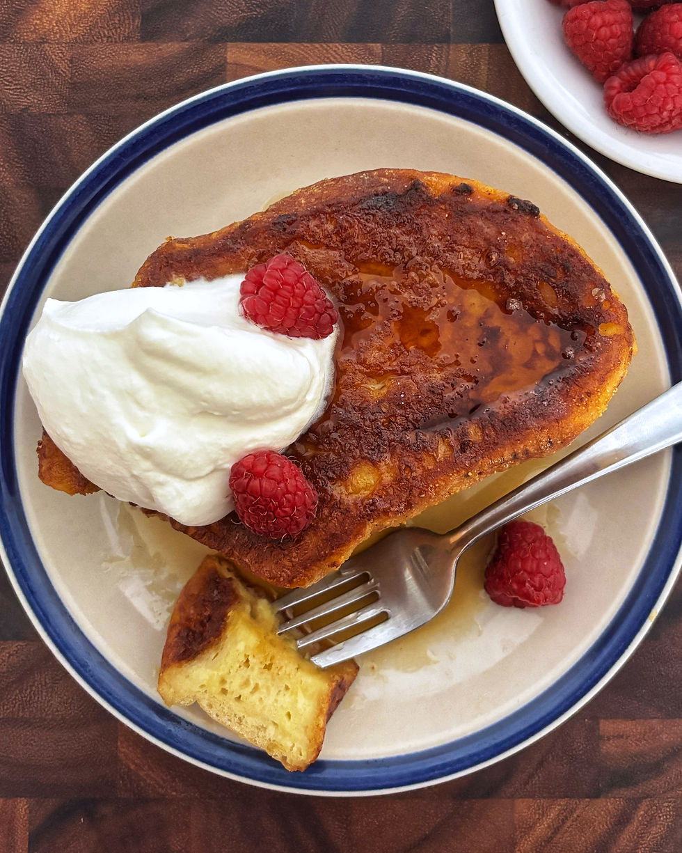 Door-stopping overnight french toast topped with whipped cream and raspberries on a plate with a fork.