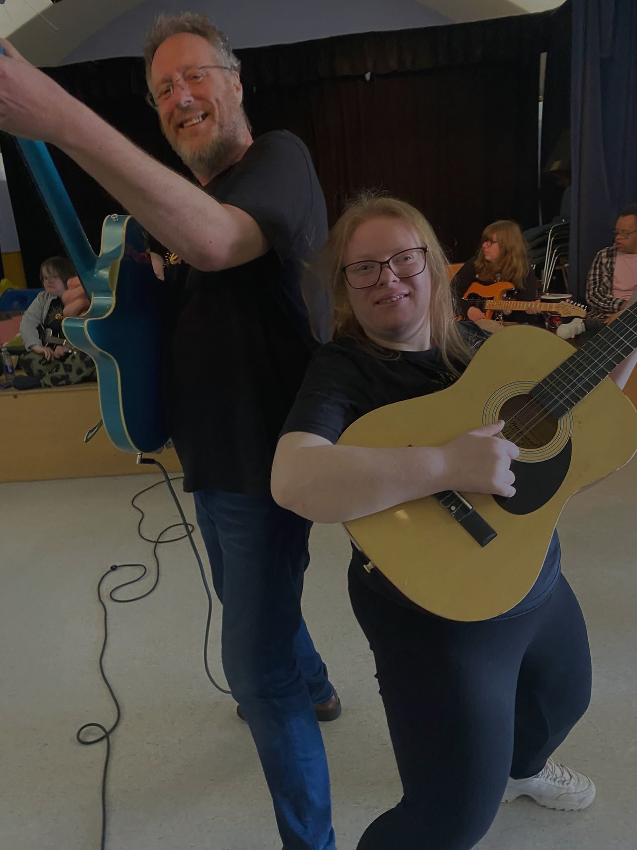 A man and a woman pose back-to-back with guitars, looking joyful. Other people with instruments can be seen in the background, which suggests a group music session.