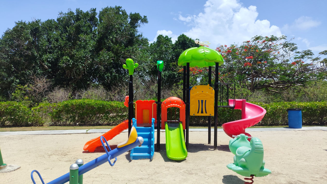 Un parque de diversiones al aire libre para los niños y adolescentes con juegos coloridos en tamaño real, asientos coloridos, toboganes y casas, al lado de una cancha de tennis y rodeado de arboles para disminuir la temperatura del día.