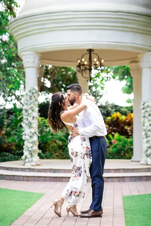 An engaged couple embraces and share a kiss in front the wedding gazebo or cupola at Villa Toscana Miami, the #1 venue for engagement photos.
