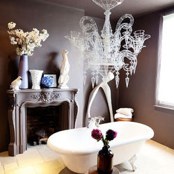 Cadă albă într-o baie sofisticată, cu pereți maro. Candelabru elaborat deasupra, statui și flori pe șemineu. Atmosferă elegantă.