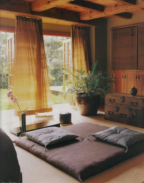 Interior calm și luminos, cu futon maro, perne gri, plante verzi și mobilier din lemn. Ferestre mari deschise către o grădină. Atmosferă zen.