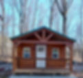 log sided cabin sitting on a pad surrounded by woods. It's front door sits on a porch with a cross beam roof and rustic railing.
