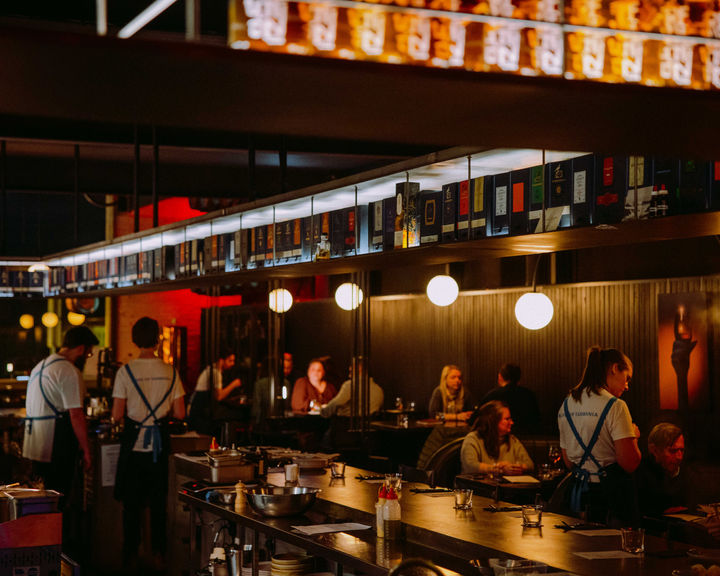 A bar full of seated people with a line of whiskies above the bar and glowing lights illuminating the bar