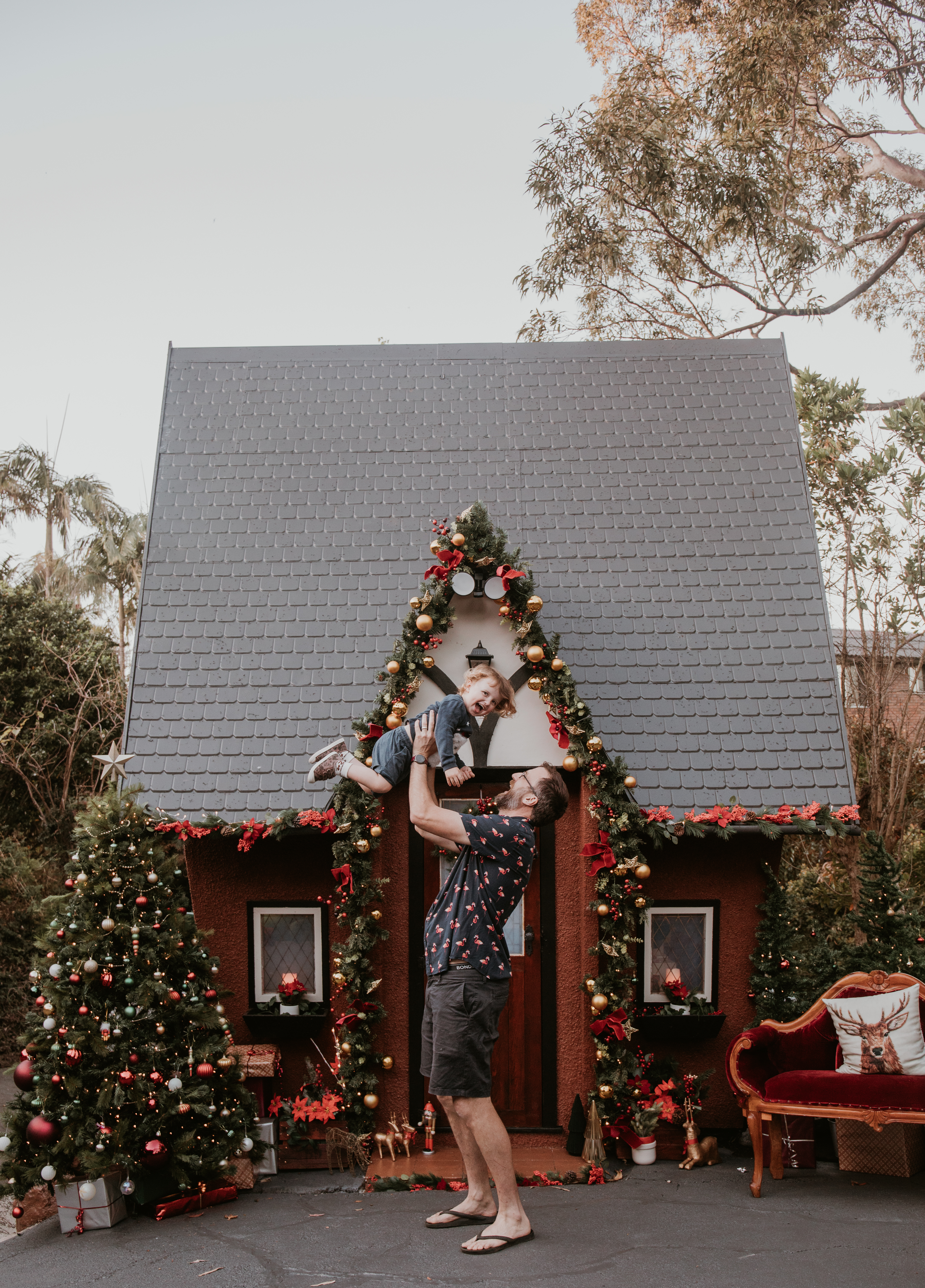 Father cuddling his young child in front of a festive Christmas Cottage decorated with greenery and red bows.