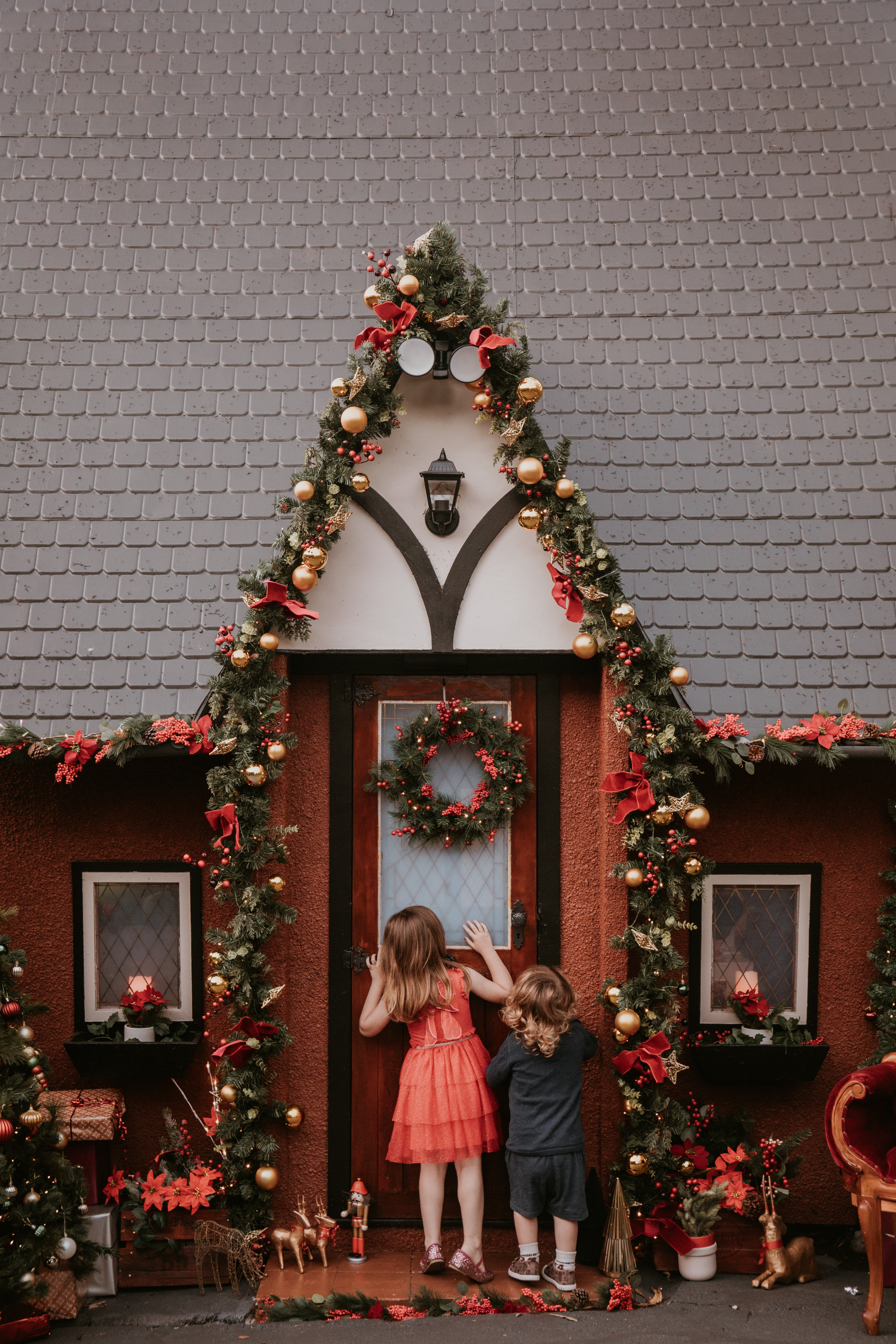 Young girl standing outside Wollongong's 
Two children exploring the decorated doorway of Wollongong's Christmas Cottage during a festive photo session. Christmas Cottage, decorated with festive garlands and Christmas lights for outdoor mini photography sessions.