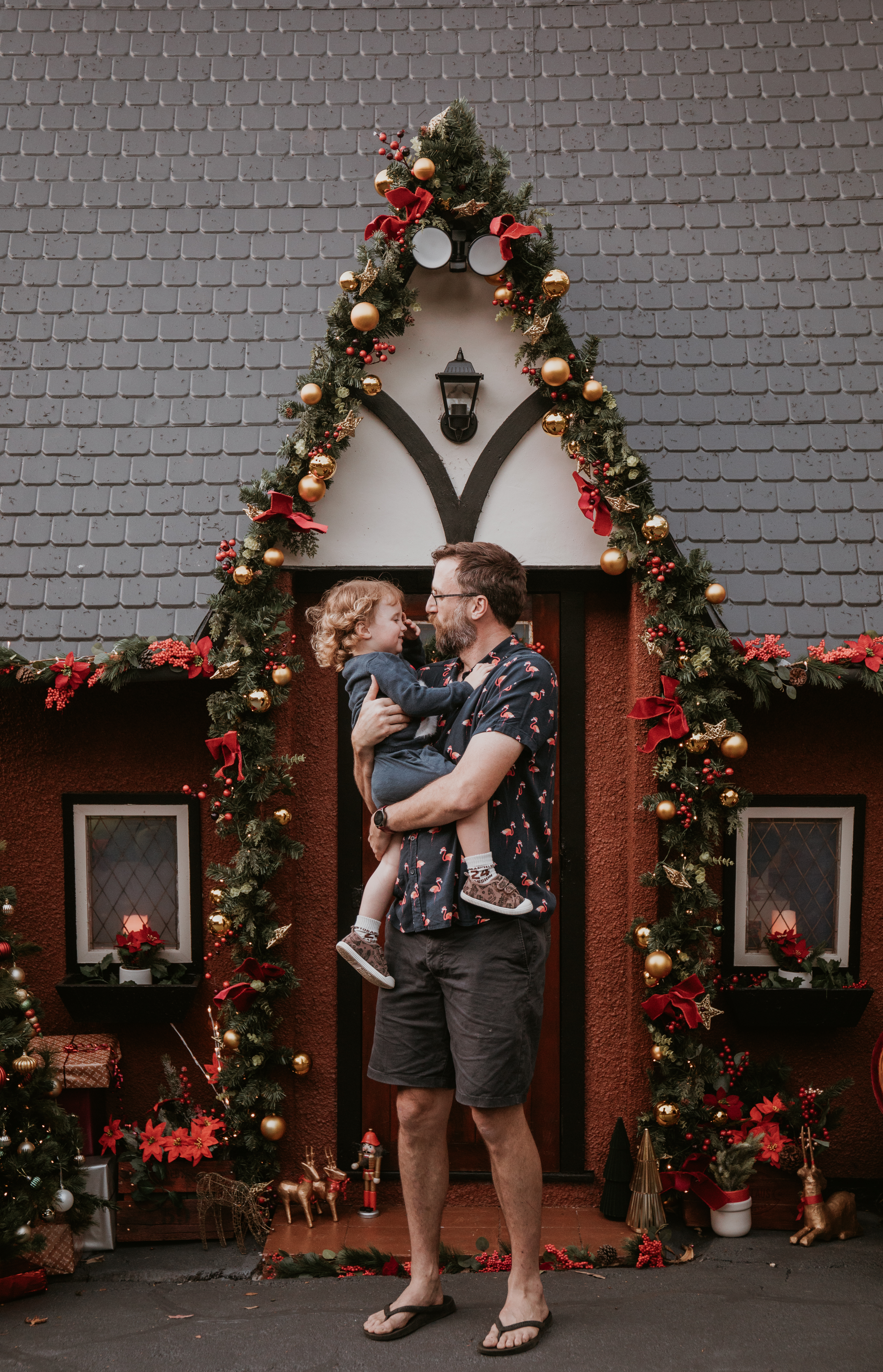 Father cuddling his young child in front of a festive Christmas Cottage decorated with greenery and red bows.