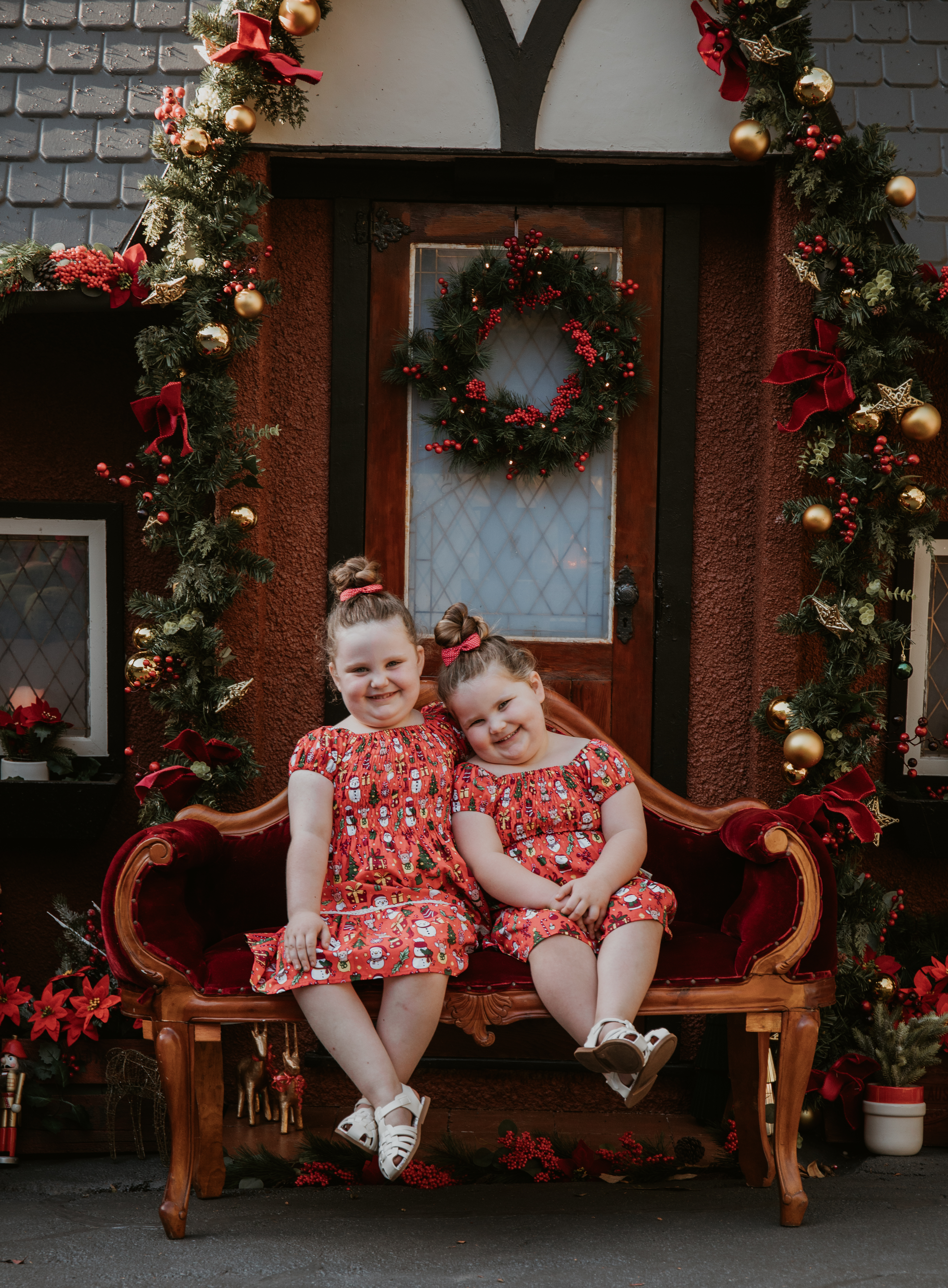 Two young girls cuddling on a vintage red velvet sofa surrounded by traditional Christmas decorations.