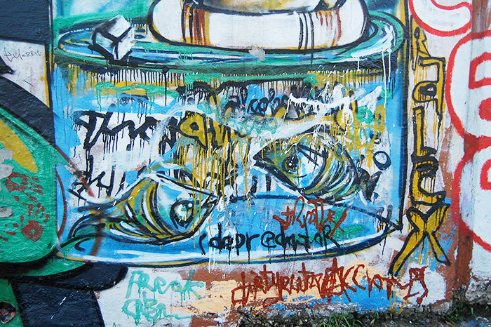 Colorful mural with abstract eyes, vibrant blue and yellow hues. Dripping paint effect. Artist's signature and graffiti text below. Urban setting.
