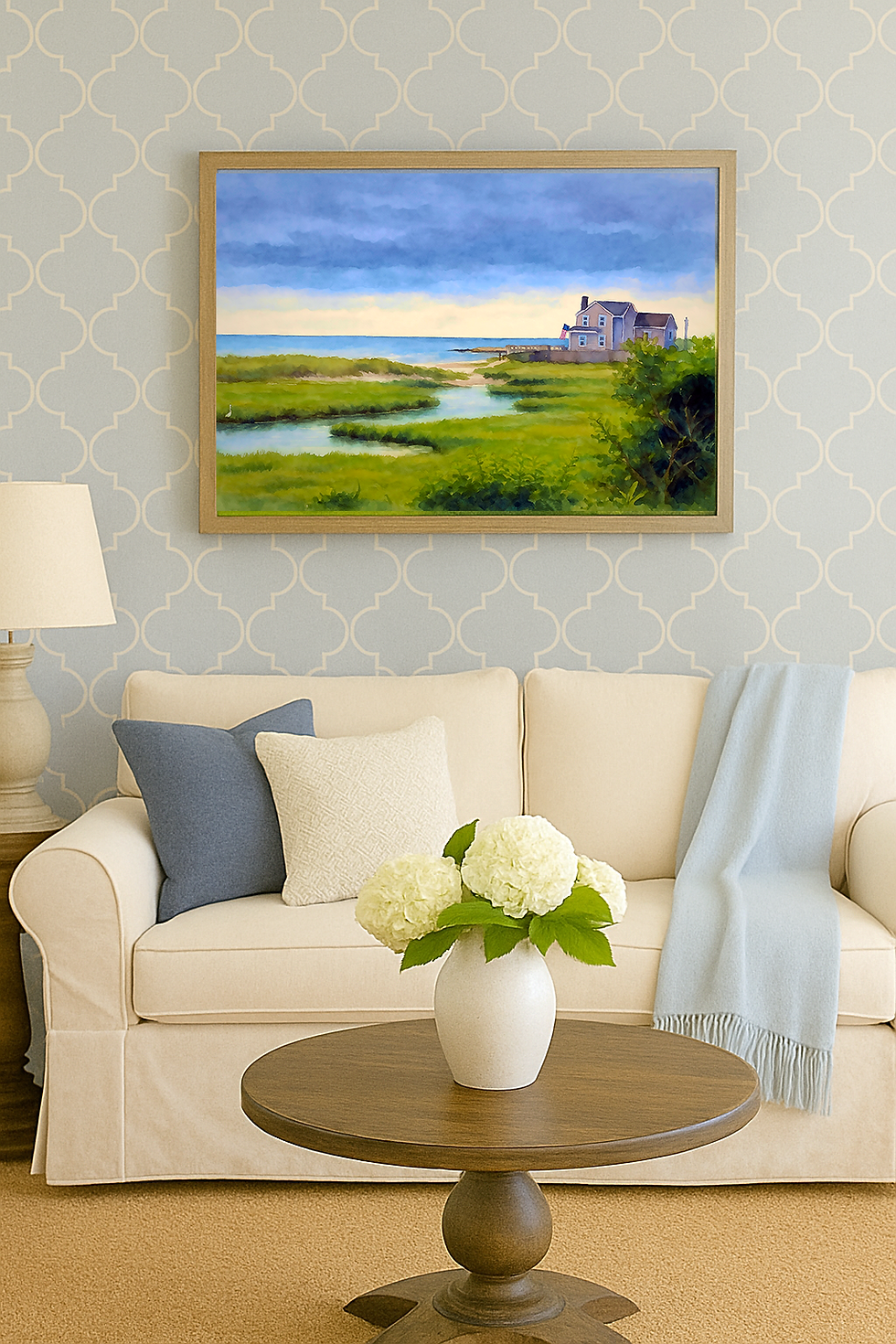 Beige sofa with blue blanket and pillows, framed marshland seascape painting with a Cape Cod Colonial house, wooden table with white vase of flowers, patterned wallpaper background.