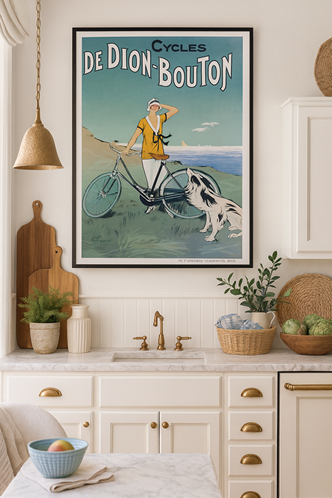 Vintage French poster of a cyclist in France with a dog by the sea, framed on a kitchen wall. White cabinets, gold brass accents, and wicker baskets nearby.