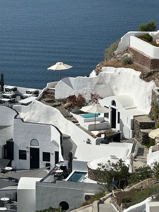 White cliffside buildings with pools overlooking the ocean, featuring a sun umbrella and blooming flowers, create a serene, sunny Santorini Greece ambiance.