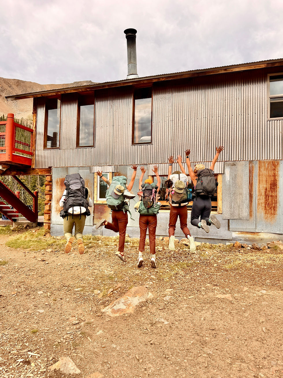 Group of women backpackers from behind jumping into the air in front of backcountry hut