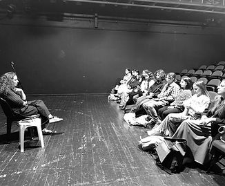 אישה יושבת על כיסא מול קהל, בחדר חשוך. ראשי, הקהל מקשיב בקשב רב.