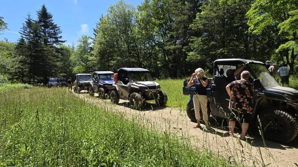 Club members and side-by-sides stopped along a wooded trail on a sunny day