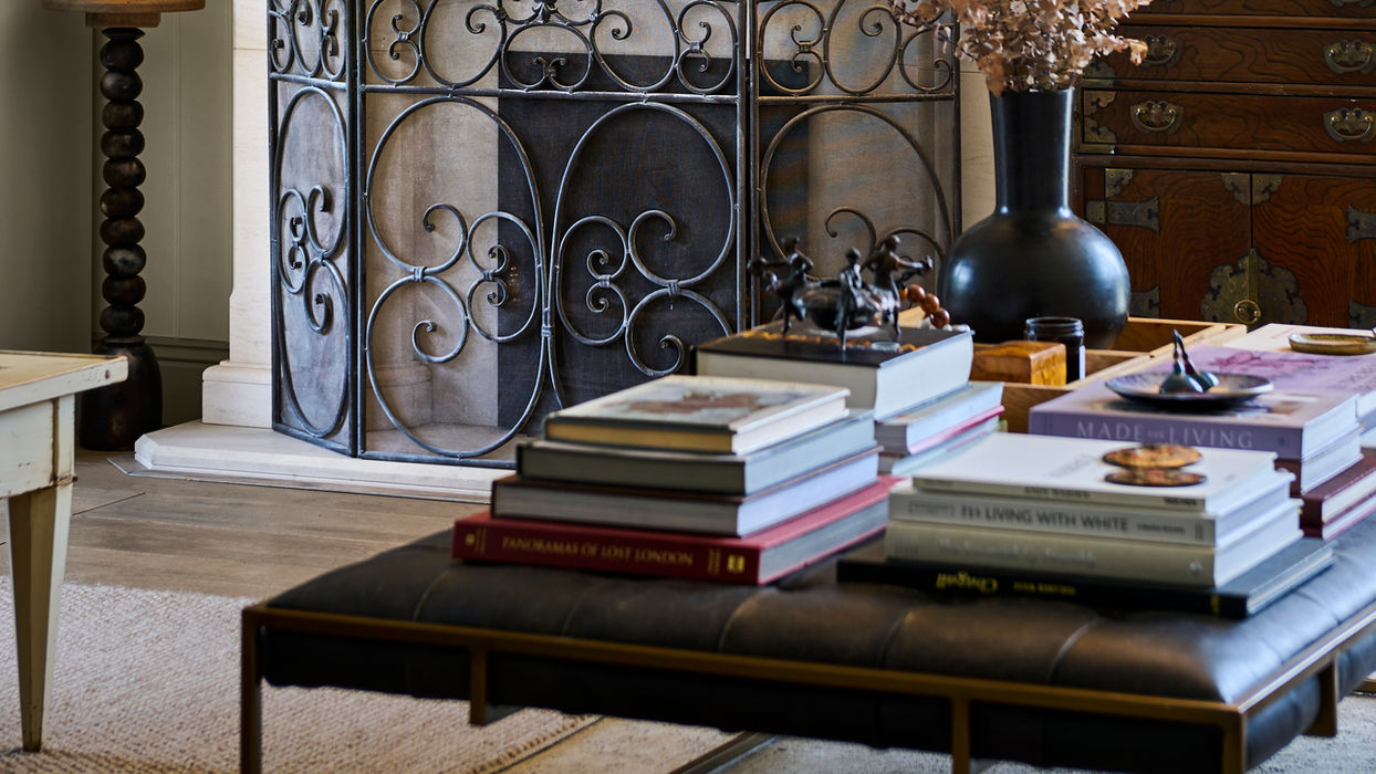 Elegant living room with limestone fireplace, round mirror, and buttoned leather ottoman covered in books.