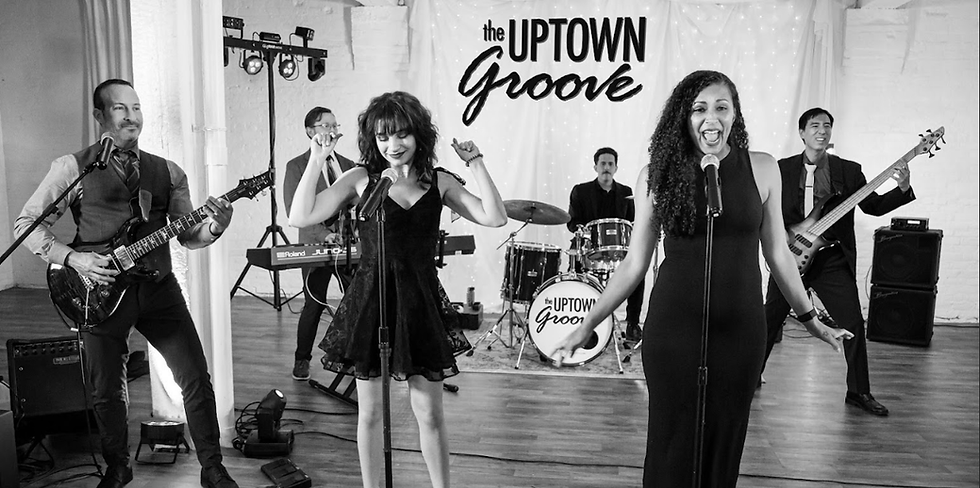 The Uptown Groove singer Taylor sings into a microphone while the rest of the band plays on stage in front of a curtain of lights.