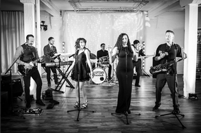 7 members of the Uptown Groove perform on stage in front of a curtain of lights.A male and female lead singer sing into microphones. The Uptown Groove logo hovers at the top of photo.