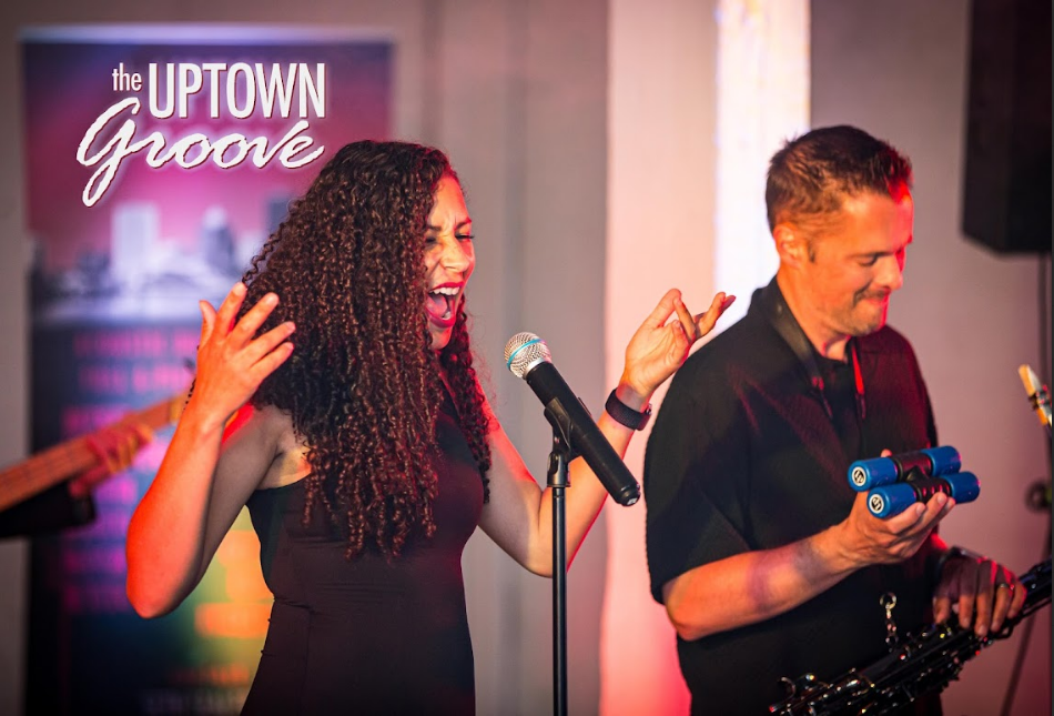 Female lead singer for The Uptown Groove sings into a microphone while a saxophonist plays auxiliary percussion next to her.