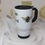 Thumbnail: Watercolour Bee and flowers with "Bee Kind" text, white Stainless Steel Travel Mug, black plastic handle & closeable lid.