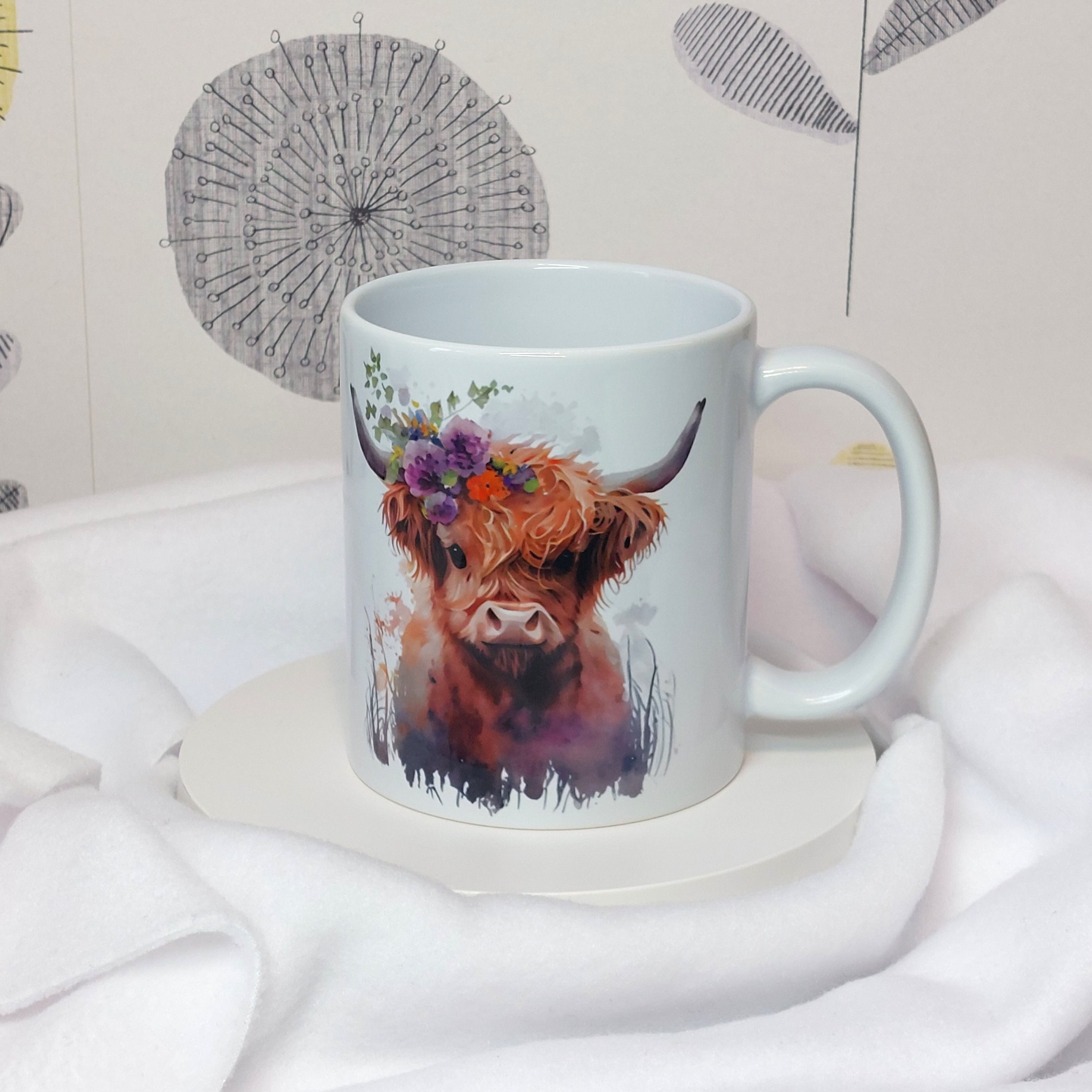 Watercolour image of a young Highland cow with flowers and botanicals on its horn with paint drips on a white mug