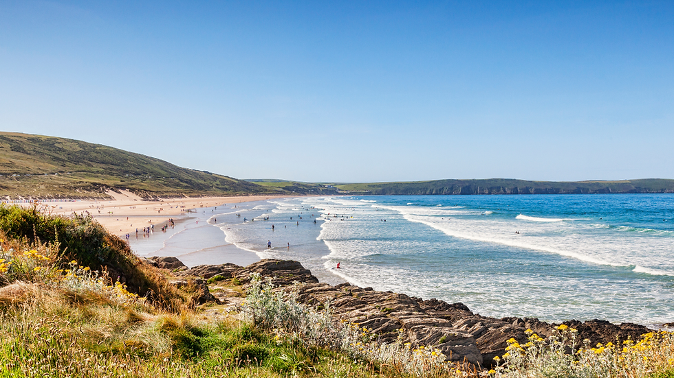 People enjoying a sunny day at a wide sandy beach in South Devon, with gentle surf and coastal cliffs in the distance.