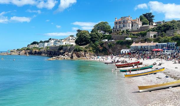 Busy summer day on Brixham’s Breakwater Beach, with colourful boats lined up along the shore and visitors enjoying the calm turquoise sea.