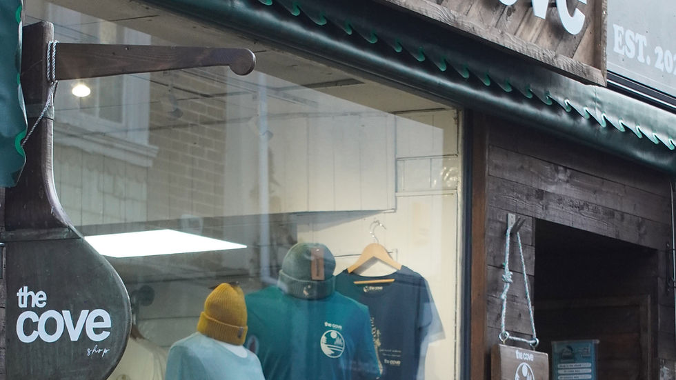 The Cove shop on Fore Street in Brixham showcases a variety of clothing items, including hats and t-shirts, enticing shoppers with its rustic wooden facade and inviting displays.