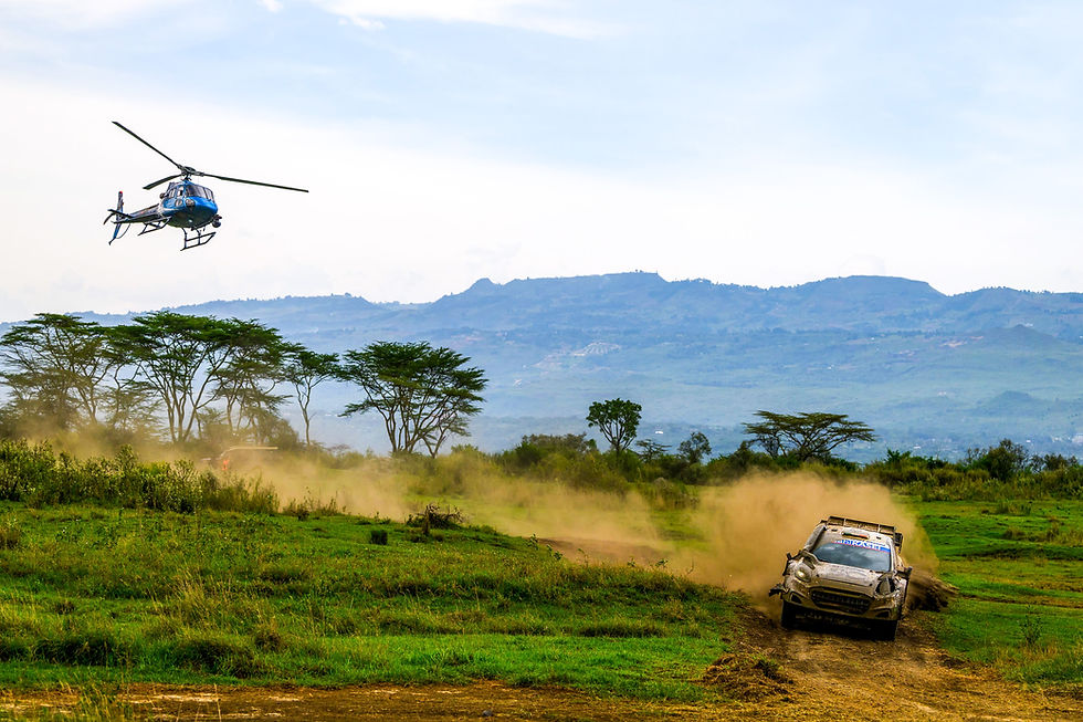 Rally car speeding on a dirt track, kicking up dust. A helicopter flies overhead. Green landscape with distant hills in the background.