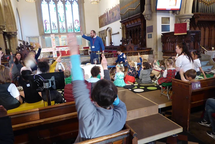 Children seated on the floor in front of the vicar at an All Age service, with hands up to answer a question.