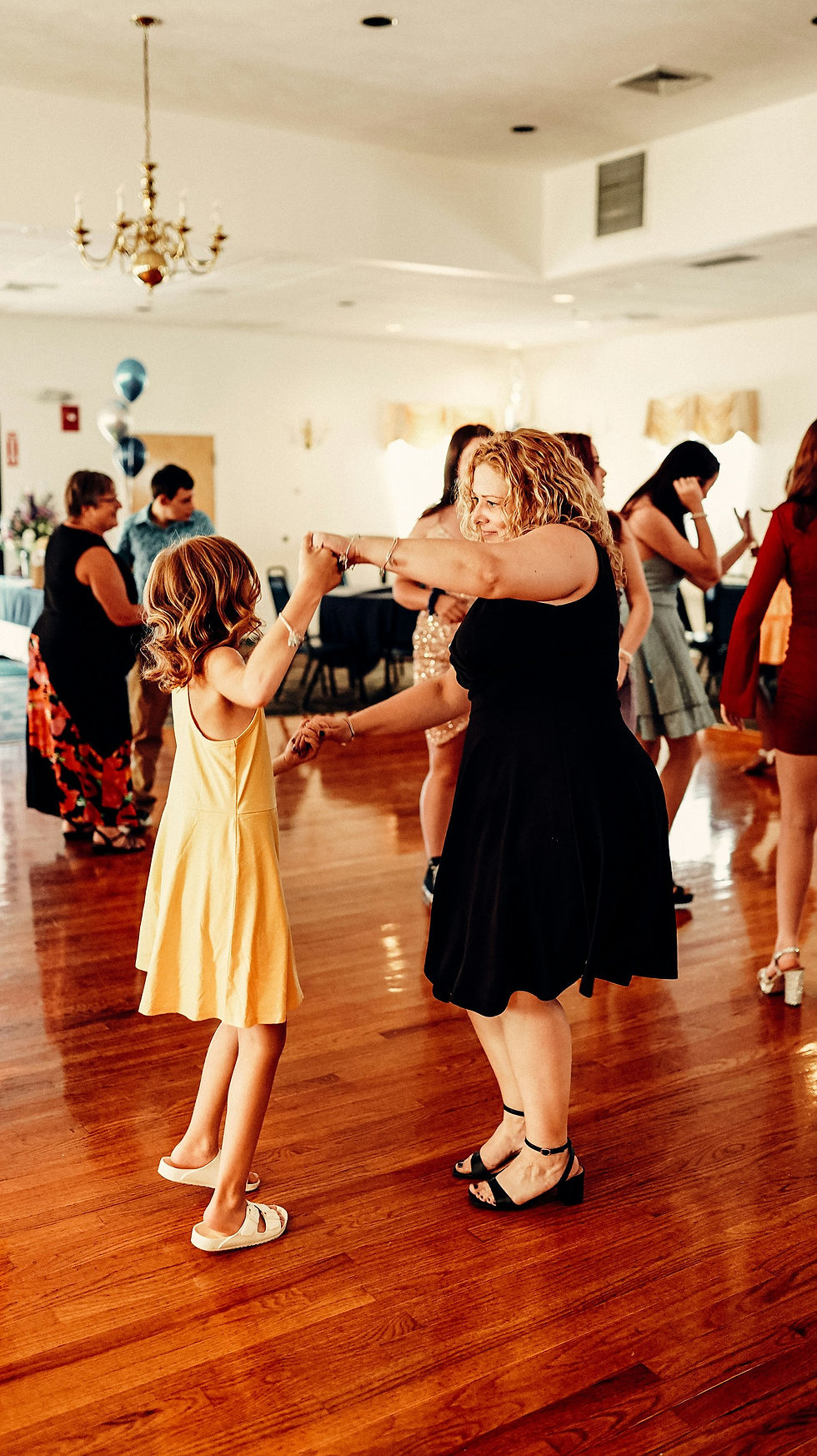 Woman in a black dress dances with a girl in yellow on a wooden floor. People enjoy a party in the background, balloons visible.