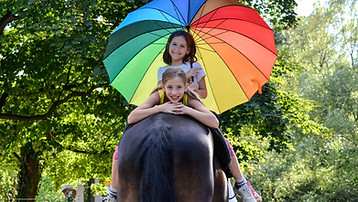 Zwei Mädchen sitzen verkehrt auf einem Pferd im Grünen und halten einen Regenbogenregenschirm