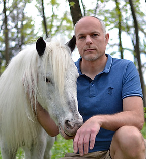 Roman Ziehengraser sitzt neben einem weißen Therapie-Pony im Wald